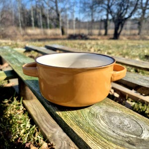 Op de afbeelding: Een mosterdgele keramische kom met twee handvatten en een donkerbruine rand. De binnenkant is wit. De kom staat op een verweerd houten oppervlak, met een vage buitenachtergrond.