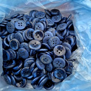 May include: A large pile of navy blue buttons with four holes each. The buttons have a marbled effect, with lighter blue swirls throughout. They are contained within a translucent blue plastic bag, suggesting they are new and unused.
