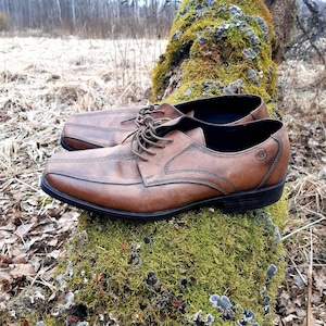 K&ouml;nnte beinhalten: Ein Paar braune Lederschn&uuml;rschuhe mit leicht abgenutztem Aussehen. Die Schuhe stehen auf einem moosbewachsenen Baumstumpf.