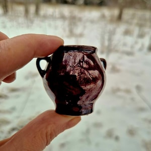 Peut inclure: Un petit pot en céramique brun foncé avec deux petites anses. Le pot a une finition brillante et est tenu entre deux doigts sur un fond flou et enneigé. Il s'agit d'un objet décoratif miniature.