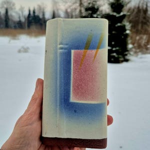Pode incluir: Vaso retangular de cerâmica com corpo creme, design geométrico azul e rosa e base marrom escuro. O design inclui duas linhas diagonais amarelas. O vaso é segurado em uma mão.