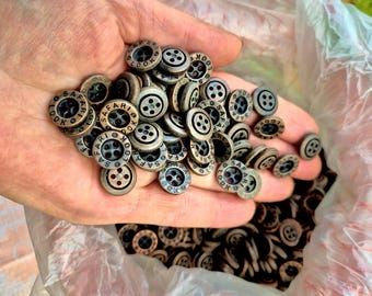 May include: A collection of bronze-colored buttons with the text "SAFARI LOOK" around the edge. The buttons have four holes and are scattered in a clear plastic bag. The buttons are small and round, with a slightly aged appearance.