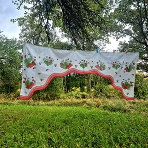 May include: A white fabric valance curtain featuring a repeating pattern of red strawberries, green leaves, and white blossoms. The bottom edge is scalloped and finished with a double red border, hanging from a clothesline with colorful clips.