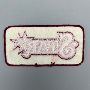 K&ouml;nnte beinhalten: Wei&szlig;er gestickter Aufn&auml;her mit einem Stern und dem Wort "STATE" auf einem maroonfarbenen Hintergrund.