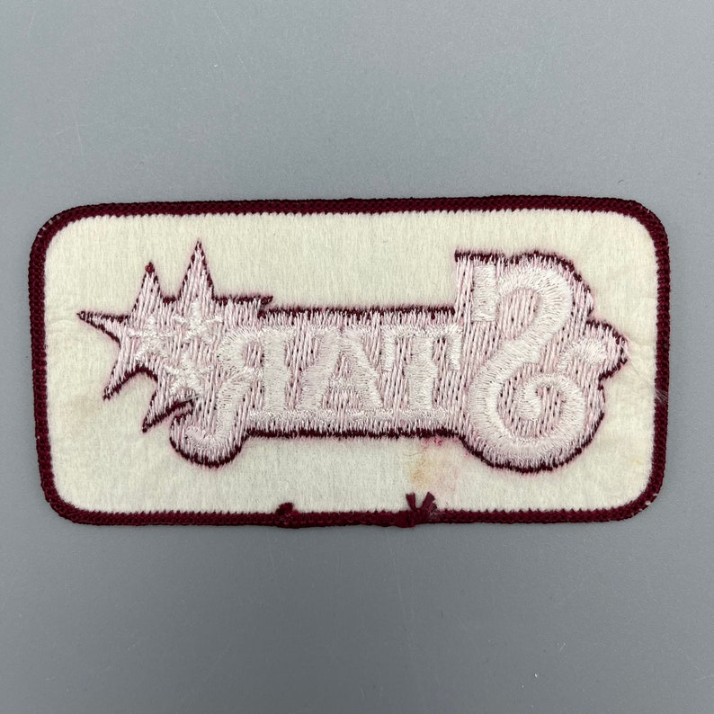 K&ouml;nnte beinhalten: Wei&szlig;er gestickter Aufn&auml;her mit einem Stern und dem Wort "STATE" auf einem maroonfarbenen Hintergrund.