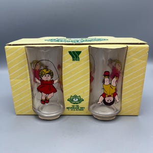 May include: A set of four 12-ounce glass beverage glasses with a Cabbage Patch Kids theme. The glasses feature illustrations of a girl with blonde hair in a red dress jumping rope and a girl with blonde hair in a yellow shirt and blue overalls doing a handstand.