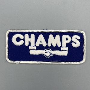 May include: A blue and white embroidered patch with the word "CHAMPS" in white lettering and a white handshake graphic.