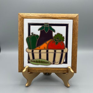 May include: A framed ceramic tile with a colorful illustration of vegetables in a basket. The vegetables include an eggplant, a green pepper, a carrot, and a tomato. The tile is on a gold stand.