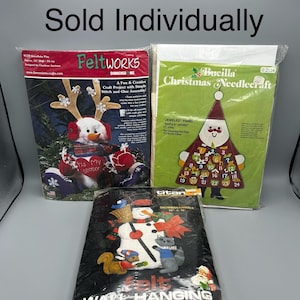 May include: Three packages of felt Christmas craft kits. The first kit features a snowman with a red hat and a reindeer antler headband. The second kit features a Santa Claus with a red hat and a green tree. The third kit features a snowman with a black hat and a blue bird. The kits are all labeled with the words "Feltworks", "Bucilla Christmas Needlecraft", and "Titan Snowman Animals".