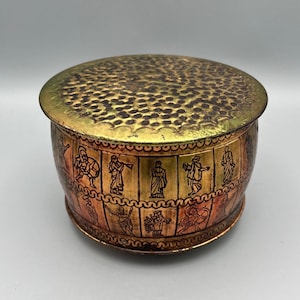 May include: A round, gold-colored metal tin with a textured lid and a band of black and red figures around the side. The figures are stylized and appear to be from a historical period.