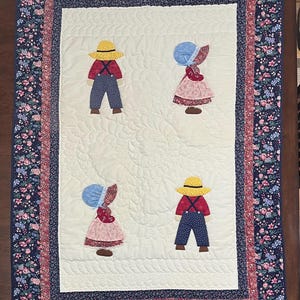 May include: Handmade quilt featuring four appliqued figures in traditional attire. The figures are dressed in red, blue, and pink clothing with straw hats and bonnets. The quilt has a floral border in shades of blue, red, and white.