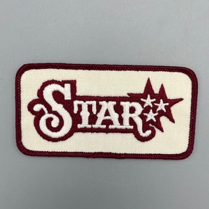 May include: A white embroidered patch with a maroon border. The patch features the word "STAR" in white with a stylized "S" in maroon. There are three white stars to the right of the word "STAR".