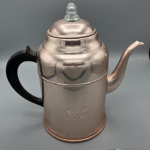 May include: A vintage copper-colored coffee pot with a black handle and a clear glass knob on the lid. The pot has a spout and a decorative emblem on the front. The coffee pot is made of metal and is likely from the mid-20th century.
