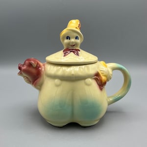 May include: A vintage ceramic teapot shaped like a person wearing overalls with a pig snout spout and a lid shaped like a person wearing a hat. The teapot is painted in shades of yellow, blue, red, and brown.