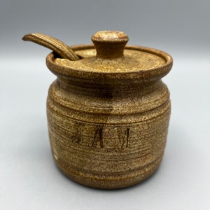 May include: A brown ceramic jar with a lid and a small spoon. The jar has the word "JAM" written on the front.