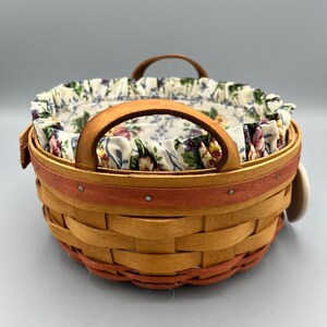 May include: A woven basket with a floral fabric liner and two handles. The basket is brown with a red rim and has a white tag attached.