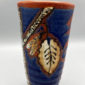 May include: A blue ceramic mug with a brown and white leaf design. The mug has a textured surface and a red rim.