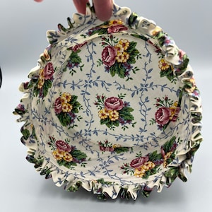 May include: A white fabric basket with a ruffled edge and a floral print featuring pink, yellow, and green roses on a white background with blue vines.