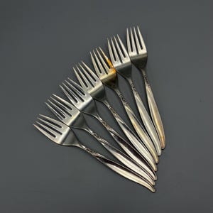 May include: Set of six silver forks with ornate designs on the handles. The forks are arranged in a fan shape.
