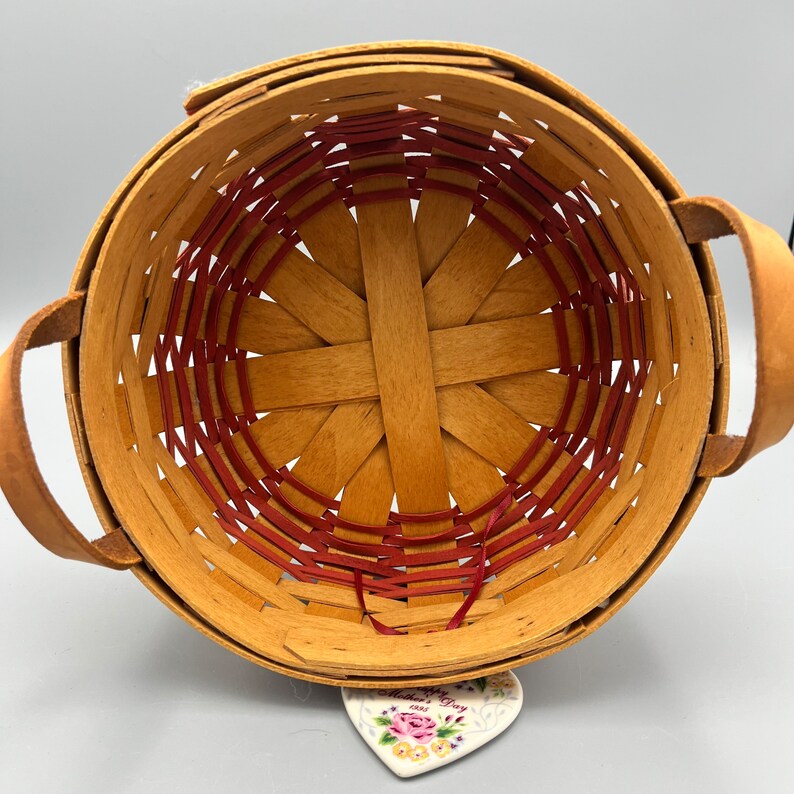 May include: A woven basket with red and brown accents. The basket has two handles and is empty. A small heart-shaped ceramic plate with a floral design and the text "Happy Mother's Day" is sitting on the bottom of the basket.