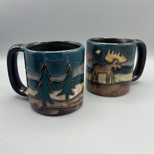 May include: Two ceramic mugs with a nature theme. One mug features two green pine trees, and the other depicts a moose with large antlers. Both mugs have a dark teal background, brown base, and black handle.