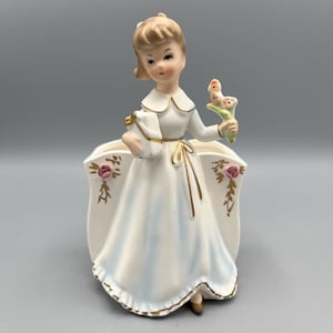 May include: A vintage porcelain figurine of a young woman in a white dress, holding a bouquet and a gift. The dress has gold accents and floral details. The figurine is a planter, with a vase-like opening at the back.