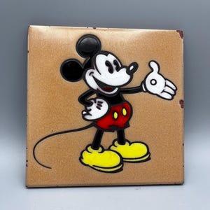 May include: A ceramic tile featuring a cartoon image of Mickey Mouse in his classic red shorts and yellow shoes. He is smiling and waving with his right hand. The tile has a tan background.