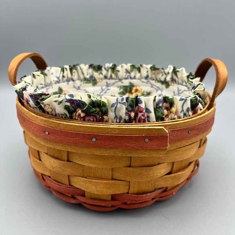May include: A small, round, woven basket with a red rim and two leather handles. The basket is lined with a white fabric with a floral pattern.