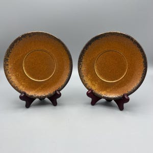 May include: Two matching ceramic saucers with a speckled, golden-brown glaze and a dark brown rim. The saucers are displayed on small, dark red stands, showcasing their circular shape and textured surface. The design is simple and elegant.