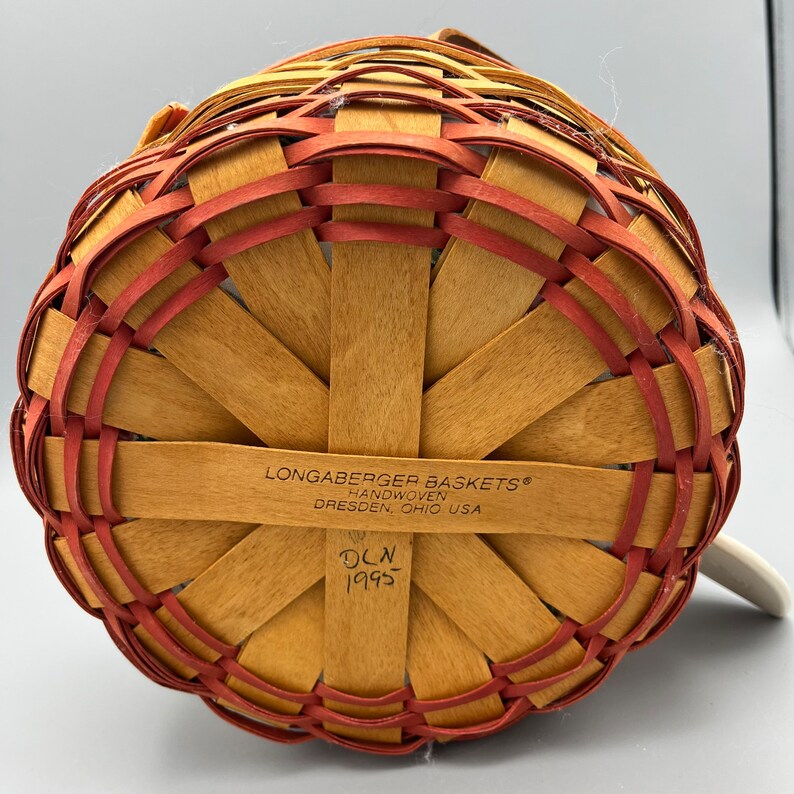 May include: The bottom of a Longaberger Baskets handwoven basket with a red and natural wood pattern. The label reads "Longaberger Baskets&reg; Handwoven Dresden, Ohio USA" and "DCLN 1995".
