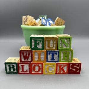 May include: Wooden alphabet blocks spelling out "FUN WITH BLOCKS". The blocks are stacked in a pyramid shape and are painted in various colors, including red, green, yellow, blue, and orange.