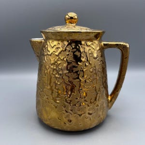 May include: A gold ceramic teapot with a textured surface and a lid. The teapot has a handle and a spout.