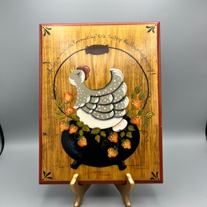 May include: A wooden plaque with a red border features a painted image of a hen sitting in a black pot with red strawberries. The text on the plaque reads "A hen a day sets the hen must lay, so to keep the stew pot full".