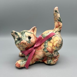 May include: A ceramic figurine of a cat with a floral pattern and a pink bow. The cat is sitting on its hind legs with its tail curled up.