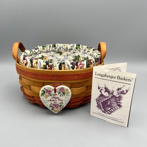 May include: A Longaberger Baskets brand woven basket with a floral fabric liner and a red and orange rim. The basket has two handles and a heart-shaped ceramic ornament with the text "Happy Mother's Day 1993". The tag reads "Longaberger Baskets - Handcrafted to be handed down."