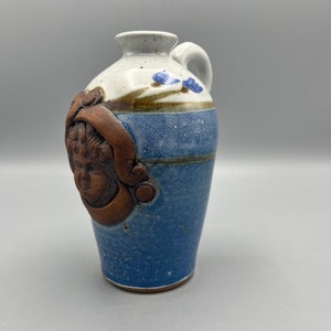 May include: A small, blue and brown ceramic pitcher with a handle and a sculpted face on the side. The pitcher has a white glaze with brown and blue accents.
