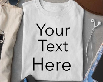 Puede incluir: Una camiseta blanca con el texto "Your Text Here" impreso en la parte delantera. La camiseta está colocada sobre una superficie gris con una cámara, un cuaderno, una bolsa de tela, un sombrero y auriculares.