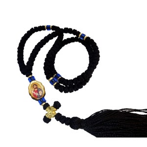 Puede incluir: Un rosario de nudos negro con un icono ovalado central que representa una figura con túnicas rojas y azules. La cuerda está decorada con pequeñas cuentas azules translúcidas y espaciadores dorados, terminando en una cruz dorada y una borla.