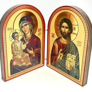 May include: A hinged religious icon featuring gold-colored illustrations of religious figures. The icon is divided into two panels, each with a detailed image. The frames are trimmed with red and gold decorative borders.