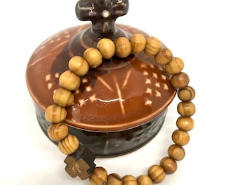 May include: A wooden rosary bracelet with a small wooden cross charm. The bracelet is resting on a brown ceramic box with a cross on the lid.