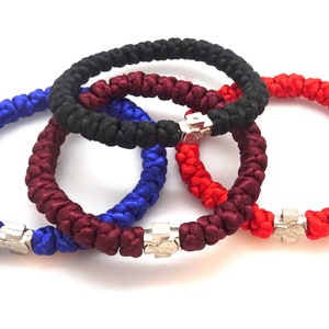 Könnte beinhalten: Vier geflochtene Armbänder mit silbernen Kreuzanhängern. Die Armbänder sind rot, blau, dunkelrot und schwarz.