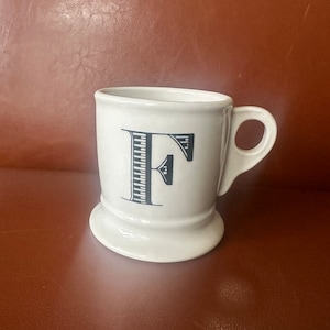 May include: White ceramic mug with a black letter F printed on the front. The mug has a wide base and a handle.