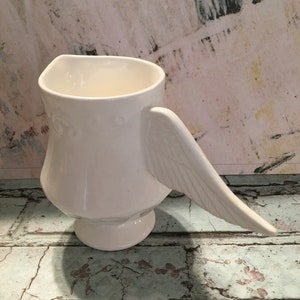 May include: White ceramic pitcher with a wing-shaped handle. The pitcher has a smooth, rounded body and a wide, open mouth.