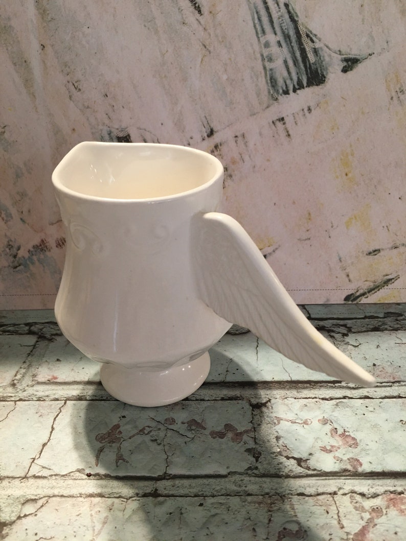 May include: White ceramic pitcher with a wing-shaped handle. The pitcher has a smooth, rounded body and a wide, open mouth.