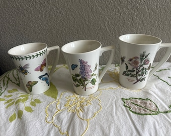 May include: Three white ceramic mugs with floral and insect designs. The mugs have a handle on the right side and a wide base.