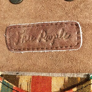 May include: Close-up of a brown leather patch with the text "Free People" embossed in white. The patch is attached to a brown suede fabric with two metal buttons.
