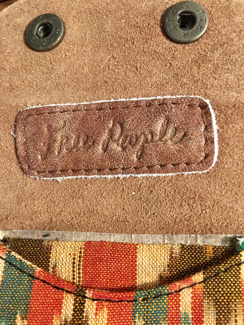 May include: Close-up of a brown leather patch with the text "Free People" embossed in white. The patch is attached to a brown suede fabric with two metal buttons.