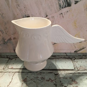 May include: White ceramic pitcher with a wing-shaped handle. The pitcher has a decorative design around the body and a round base.
