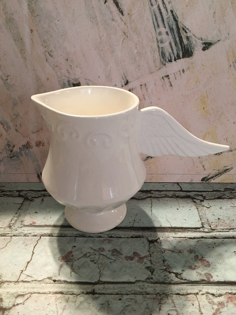 May include: White ceramic pitcher with a wing-shaped handle. The pitcher has a decorative design around the body and a round base.