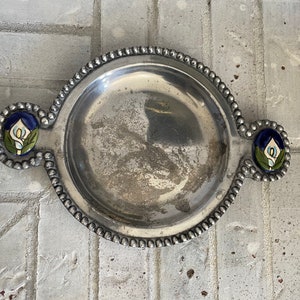 May include: A silver serving dish with a beaded rim and two floral accents. The dish is round with a slightly raised edge and has a smooth, metallic finish.